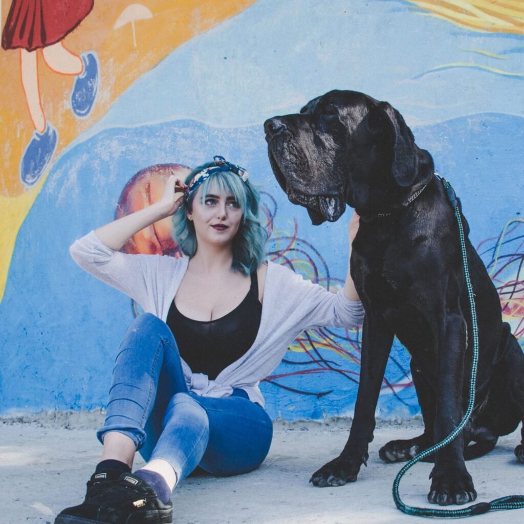 A person with blue hair sits on the ground, smiling, next to a large black dog. They are in front of a colorful mural that includes the legs of a person walking above them. The person is wearing a white cardigan and jeans.